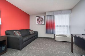 A modern hotel room with a black leather sofa, a small desk, and a large window with sheer curtains. - Ramada by Wyndham Sioux Falls Arpt & Suites (Sioux Falls)
