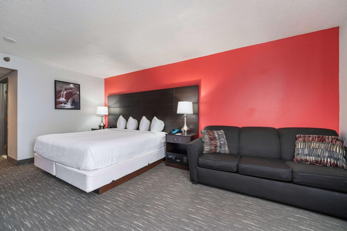 A hotel room with a large bed, a black sofa, a red accent wall, and a nightstand with lamps.