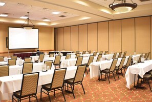 A conference room with rows of tables and chairs, a projector screen, and a podium. - Ramada by Wyndham Sioux Falls Arpt & Suites (Sioux Falls)