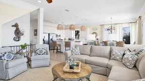 A spacious living room with a large beige sectional sofa, grey armchairs, and a round coffee table. A kitchen with white cabinets and a dining area with wicker chairs is visible in the background.