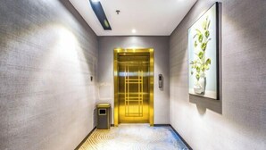 Ein Flur mit strukturierter Wand, einer gerahmten Pflanze, einem Mülleimer und einer goldenen Aufzugstür.