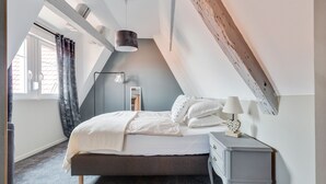 Un dormitorio con techo inclinado, una cama con ropa de cama blanca, una mesita de noche con lámpara y una lámpara de pie.