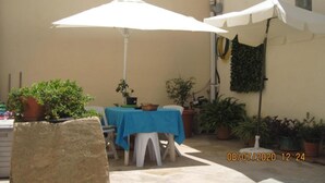A patio area with a table covered by a blue tablecloth, surrounded by potted plants, and two white umbrellas providing shade.