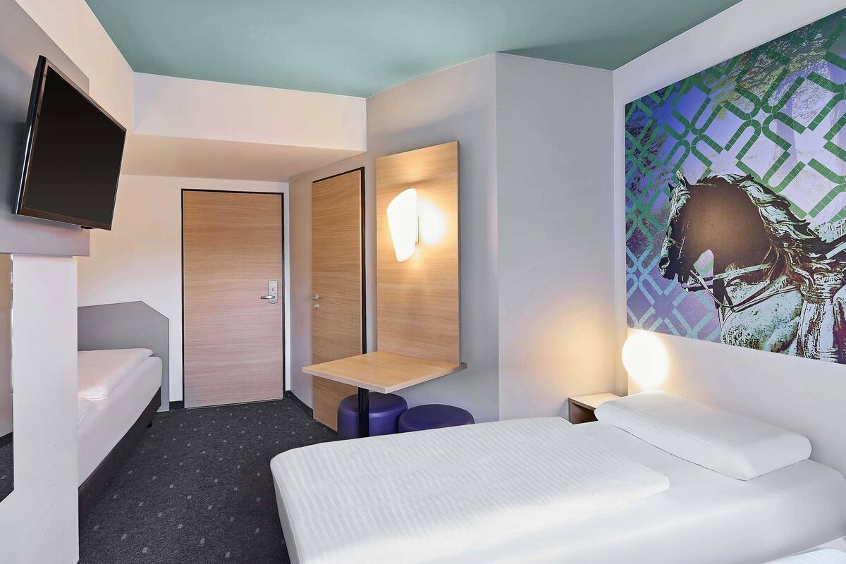 Una habitación de hotel moderna con cama, televisión montada en la pared, puerta de madera, espejo y lámpara fijada a la pared.
