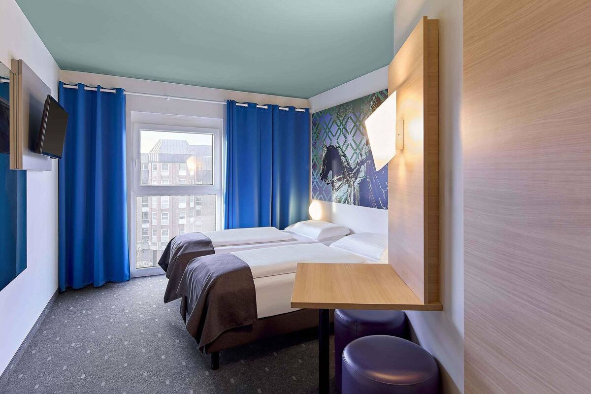 Una habitación de hotel con una cama, dos mesitas de noche, una ventana con cortinas, vistas a la ciudad y un televisor montado en la pared.