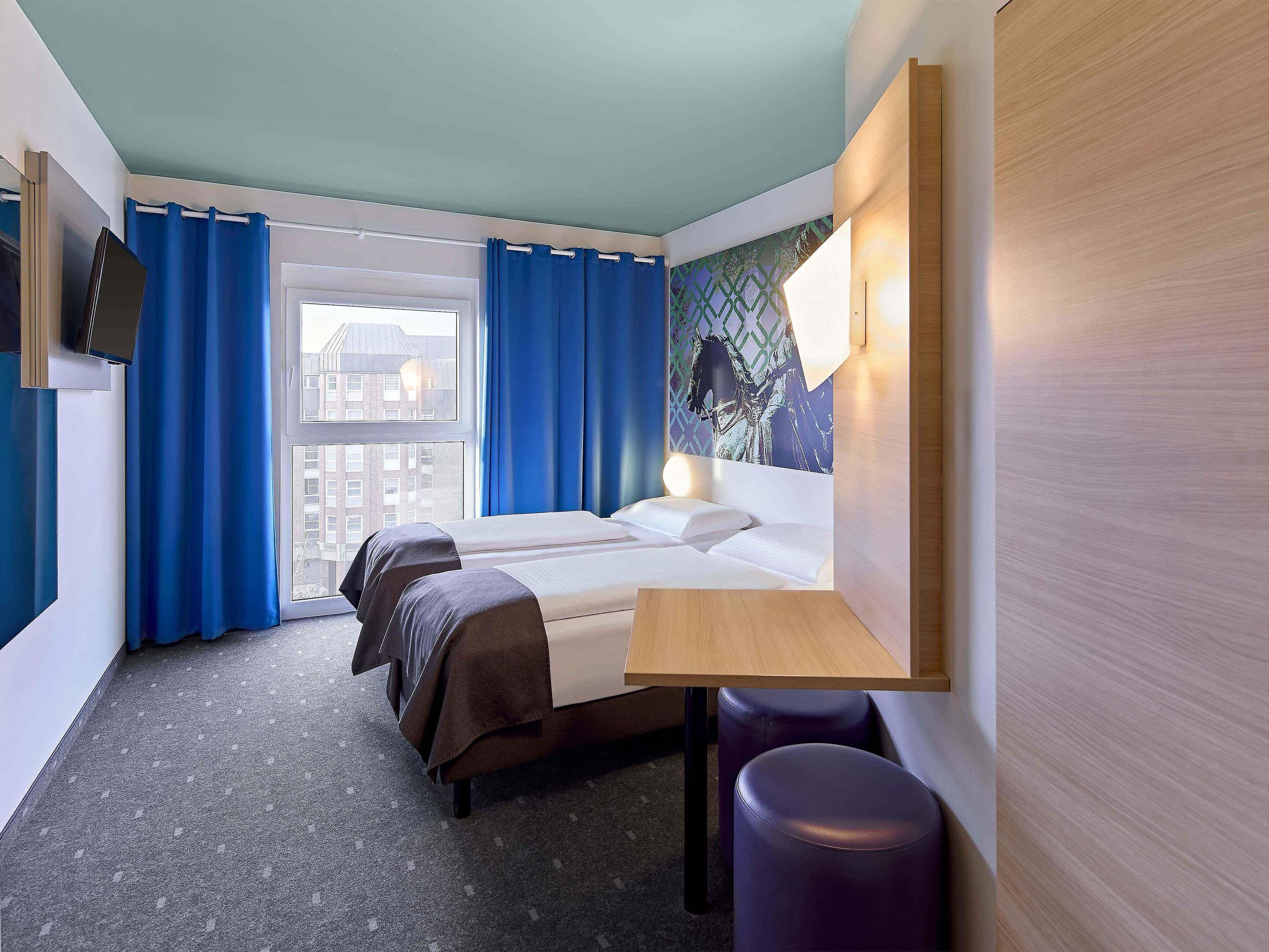 Una camera d'albergo con un letto, due comodini, una finestra con tende e televisione a parete, con vista sulla città.
