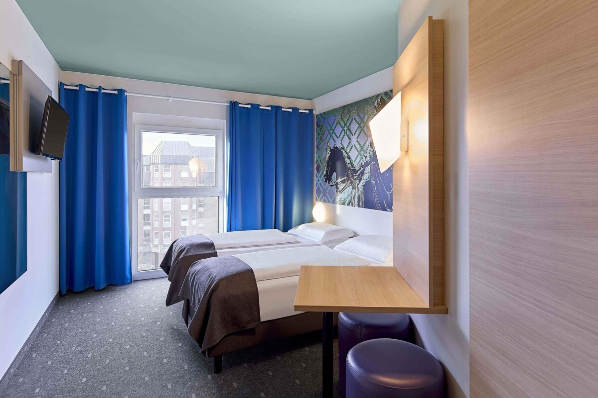 Una habitación de hotel con una cama, dos mesitas de noche, una ventana con cortinas, vistas a la ciudad y un televisor montado en la pared.