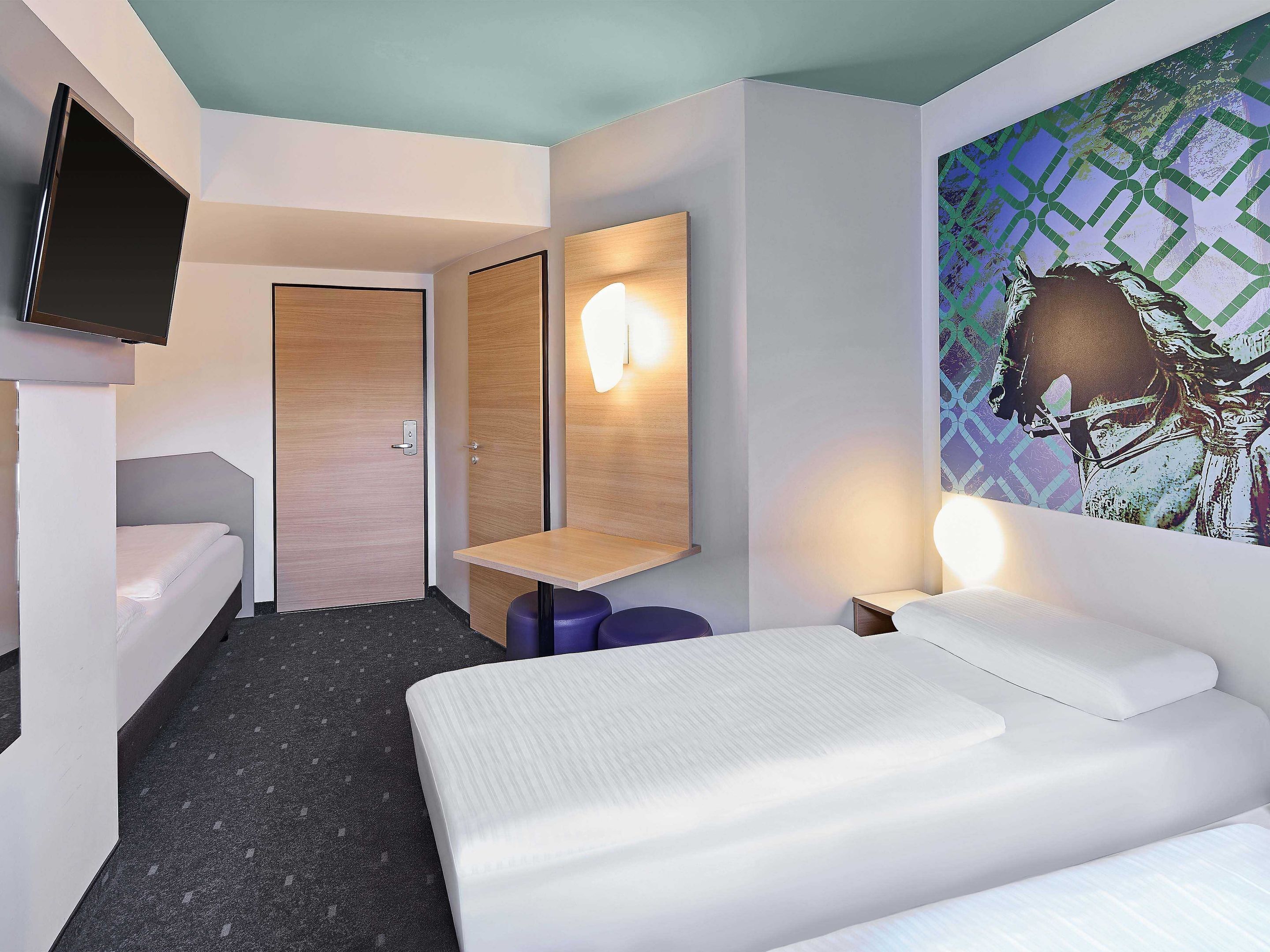 A modern hotel room with a bed, a TV mounted on the wall, a wooden door, a mirror, and a wall-mounted light fixture.