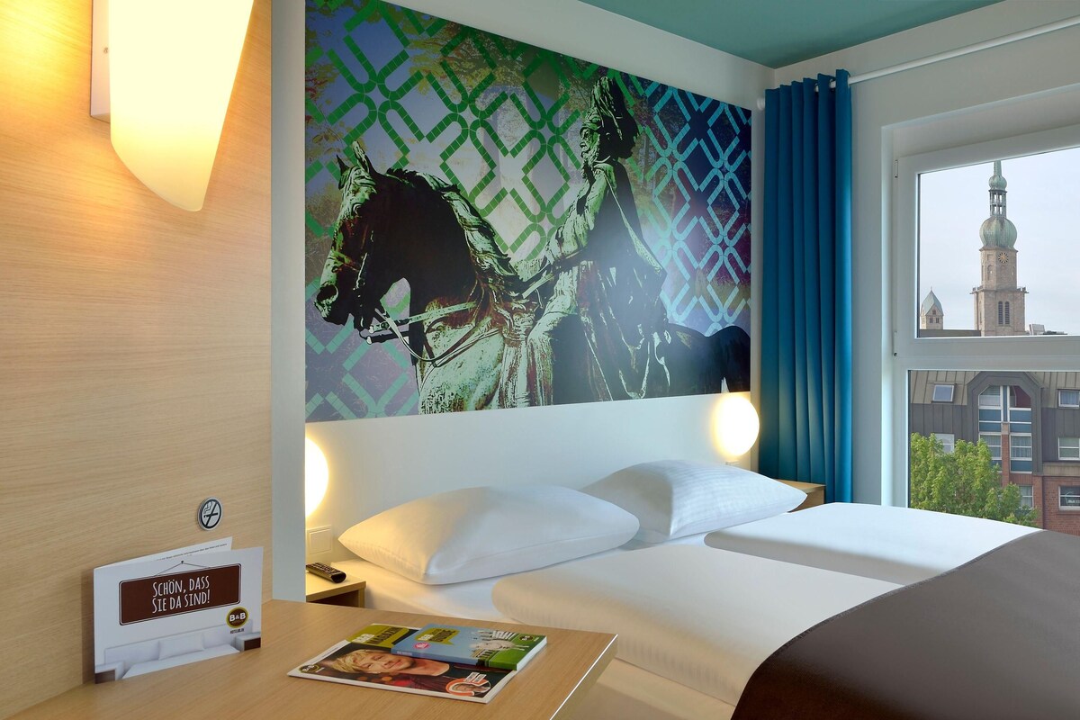 Habitación de hotel con cama, mesita de noche, lámpara, cuadro de un caballo, ventana con vista a un edificio y un cartel en la pared.