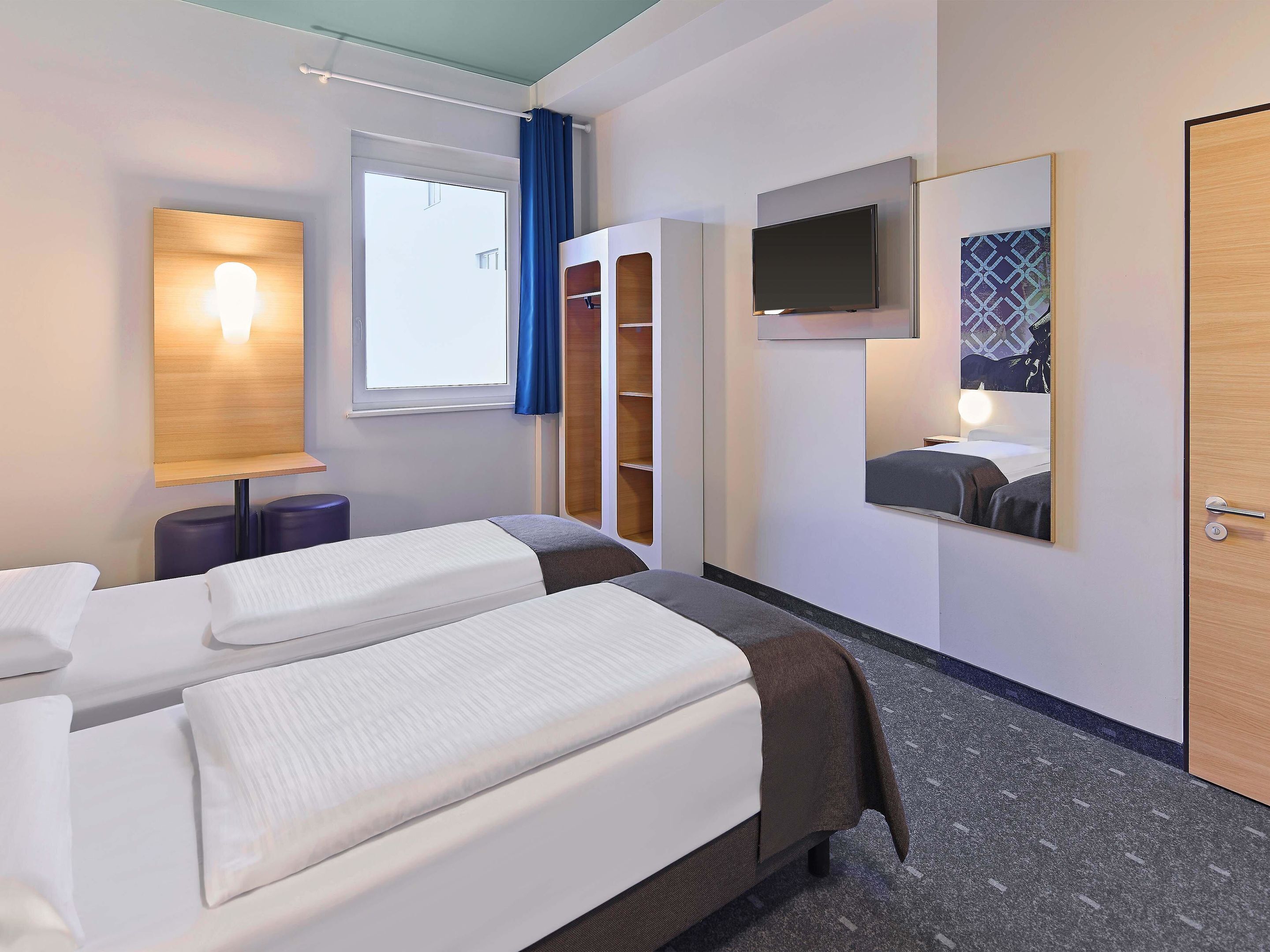 A hotel room with two beds, a TV, a mirror, a window with curtains, and a wardrobe.