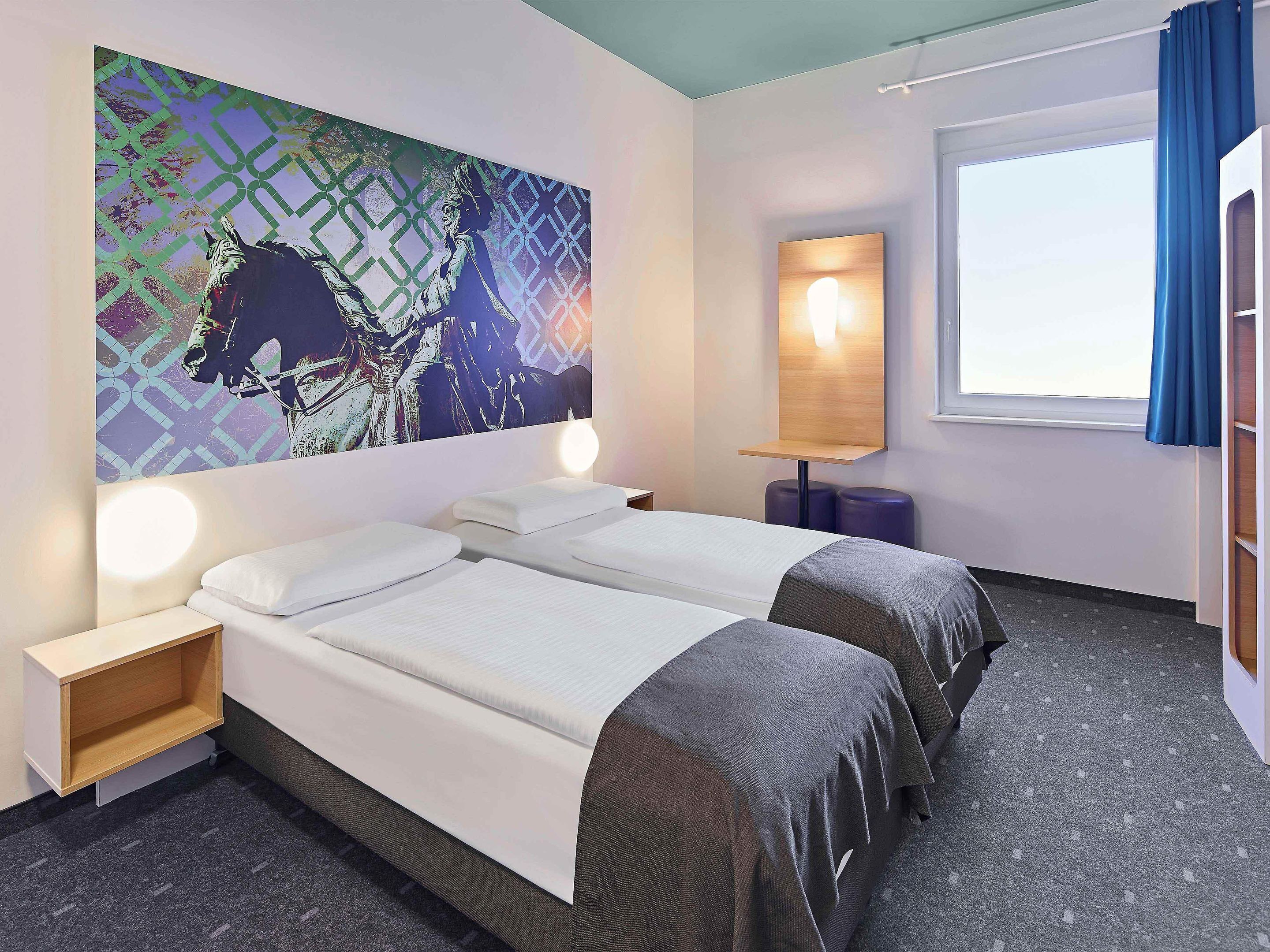 A hotel room with two beds, a large abstract painting on the wall, a bedside table with a lamp, and a window with curtains.