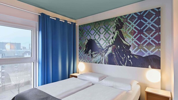 Een hotelkamer met een bed, twee nachtkastjes met lampen, een raam met blauwe gordijnen en uitzicht op de stad.
