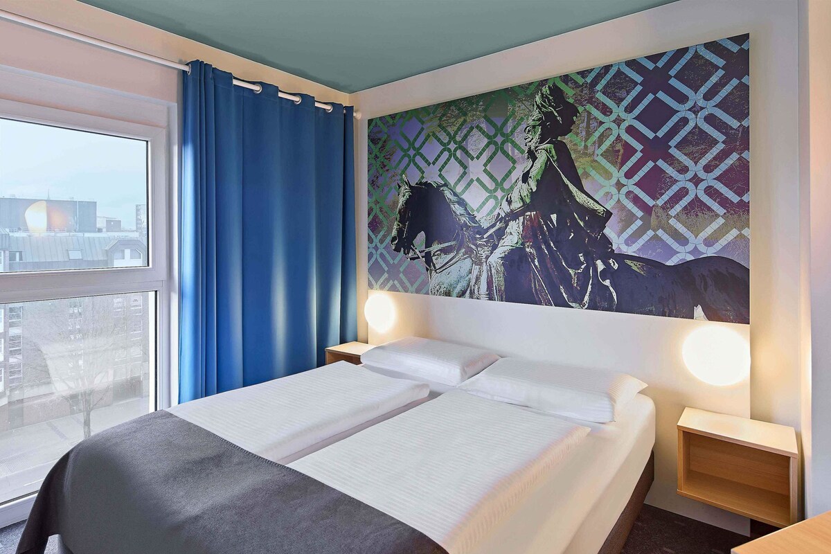 Una habitación de hotel con cama, dos mesitas de noche con lámparas, una ventana con cortinas azules y vistas a la ciudad.