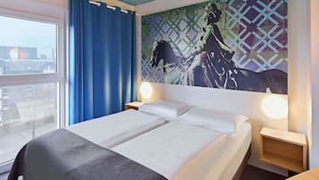 A hotel room with a bed, two bedside tables with lamps, a window with blue curtains, and a cityscape view.