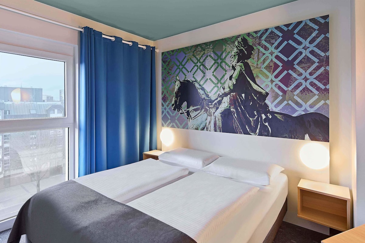 Una habitación de hotel con cama, dos mesitas de noche con lámparas, una ventana con cortinas azules y vistas a la ciudad.