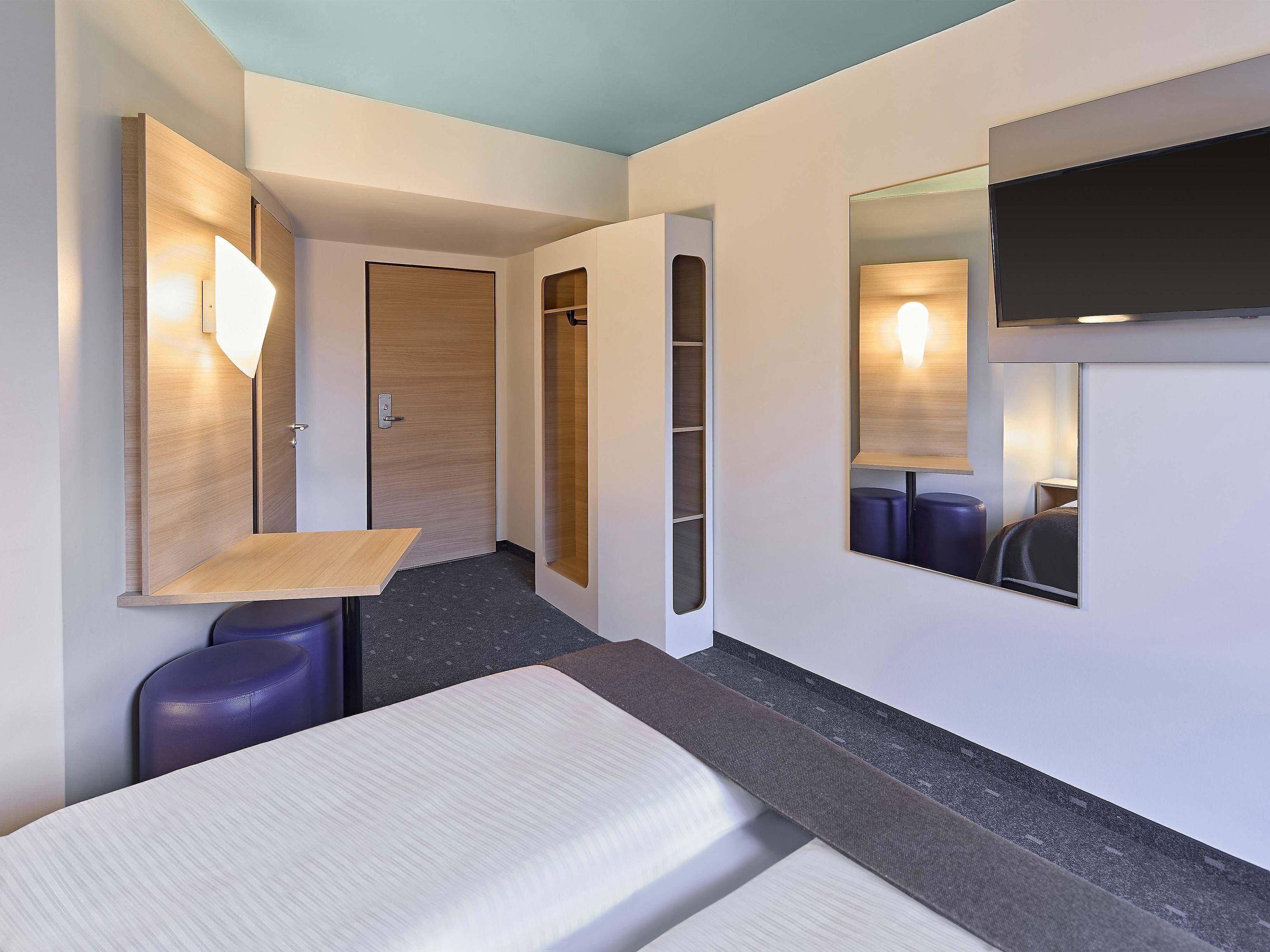 Una habitación de hotel moderna con una cama, un pequeño escritorio, un espejo y un armario.