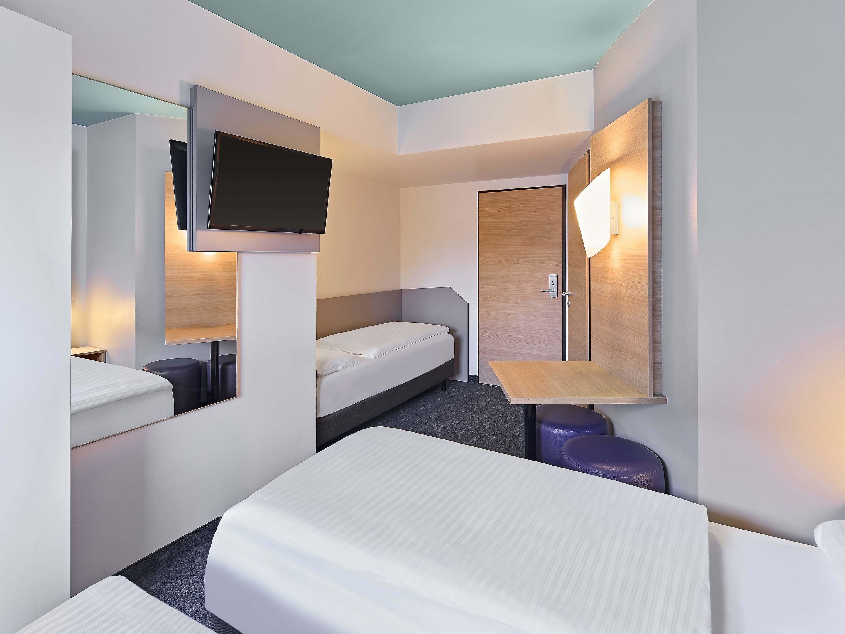 A modern hotel room with a bed, a desk, a chair, a TV mounted on the wall, and a door with a light above it.