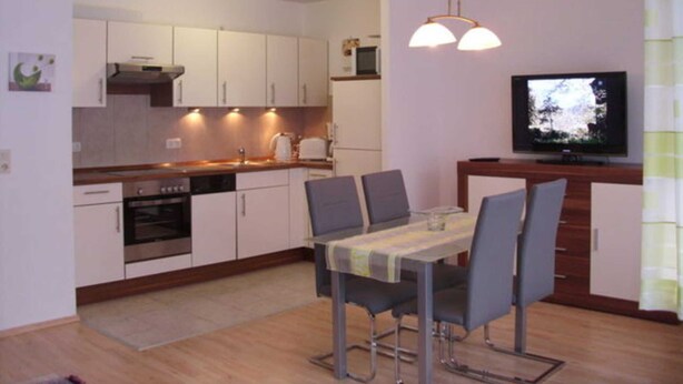 Cocina compacta con armarios blancos, un microondas y una estufa. Hay una mesa de comedor con sillas y un televisor en la pared.