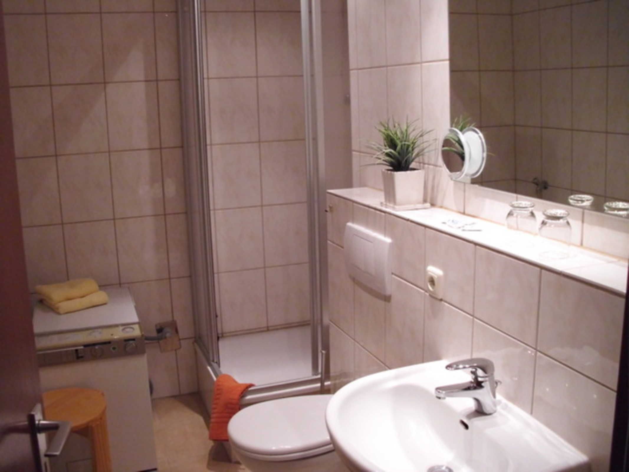 Casa de banho com sanita, lavatório e chuveiro. Há um banquinho pequeno e um vaso de plantas no balcão.