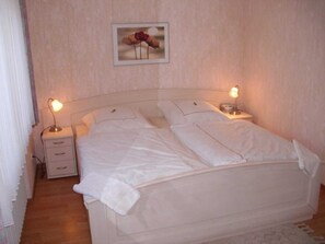Ein ordentlich bezogenes Bett mit weißen Leinentüchern, zwei Nachttische mit Lampen und ein gerahmtes Bild an der Wand.