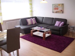 Ein modernes Wohnzimmer mit einem dunkelgrauen Sofa, einem Glastisch und einem violetten Teppich.