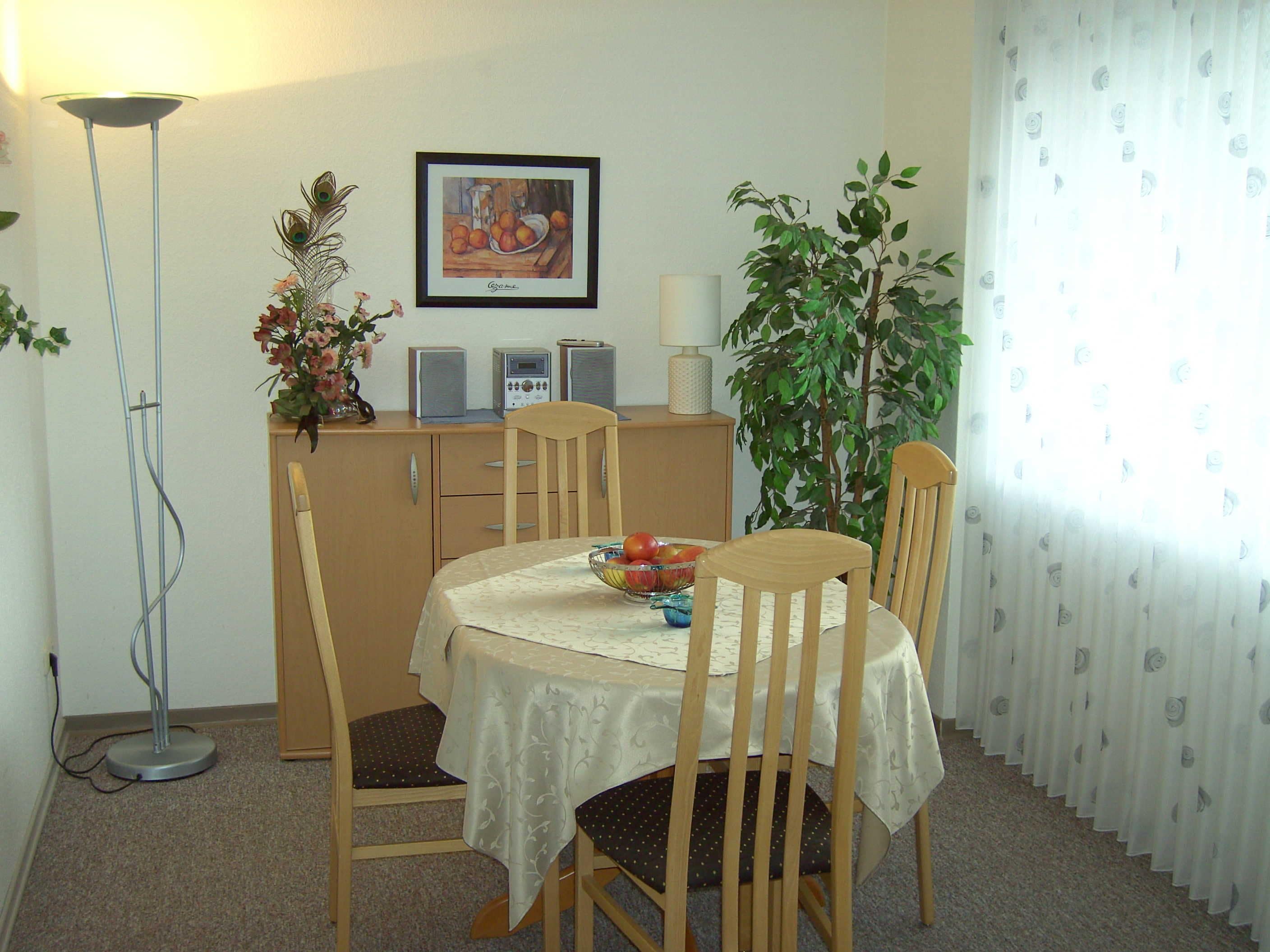 A dining area with a table set for four, surrounded by wooden chairs. There is a plant on the right and a framed artwork on the wall.