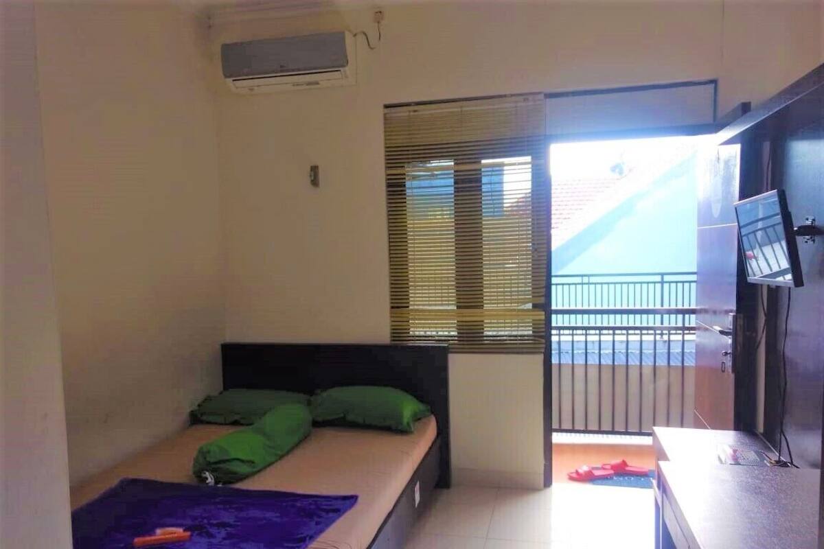 A small, well-lit room with a bed, a desk, and a window with blinds.