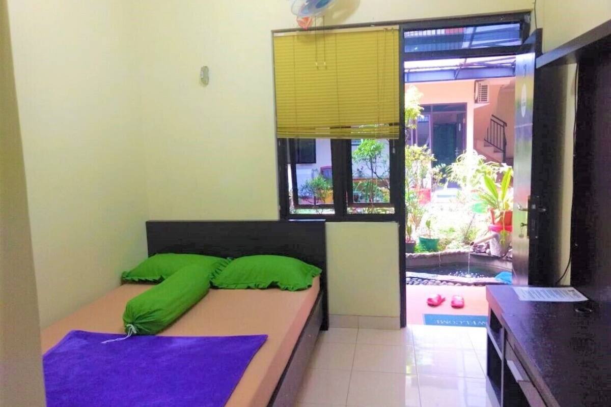 A small room with a bed, a fan, a window with blinds, and a view of a courtyard.