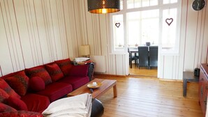 Un salon chaleureux avec un canapé rouge, une table basse en bois et une lampe. Une fenêtre est décorée d’un motif en forme de cœur et offre une vue sur un coin repas.