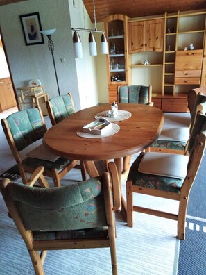 Uma mesa de jantar de madeira cercada por cadeiras com almofadas estampadas, um armário de madeira com prateleiras e um lustre.