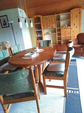 A cozy living room with a wooden dining table, matching chairs, and a sofa. There is a built-in wooden cabinet with shelves and a ladder.
