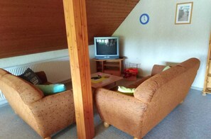 Ein gemütliches Wohnzimmer mit Holzbalkendecke, einem Fernseher auf einem Lowboard, zwei Sofas und einem kleinen Tisch mit Gegenständen darauf.