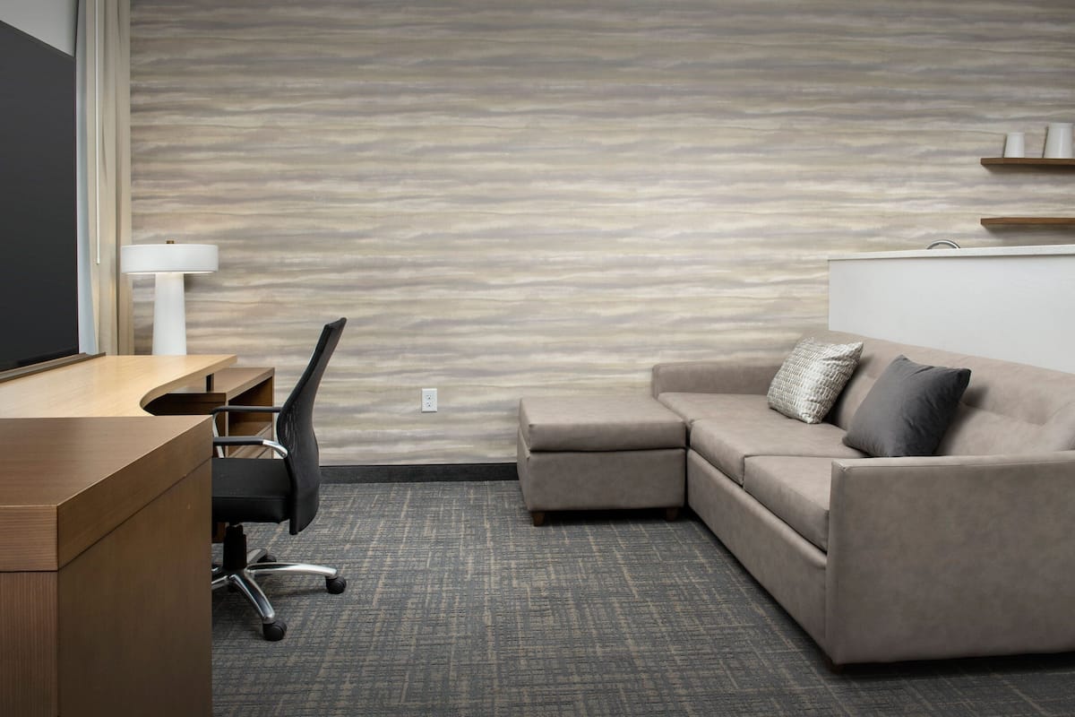 A modern hotel room with a wooden desk, black office chair, beige sofa, and a patterned carpet.