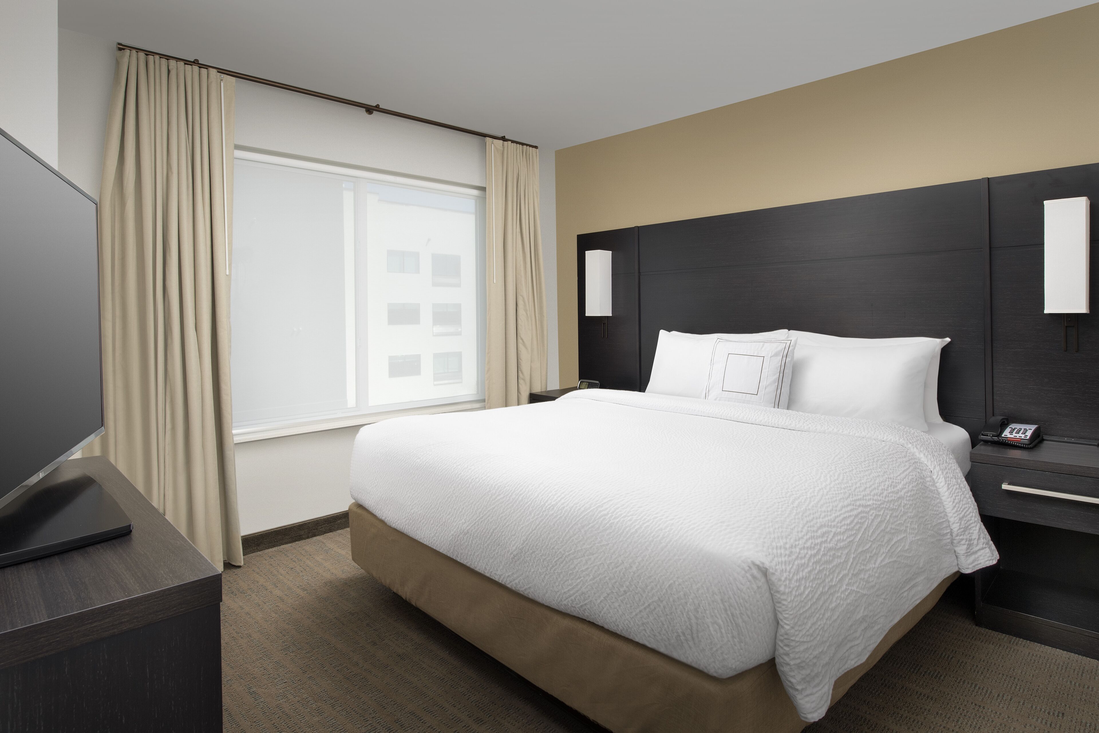 Una habitación de hotel con una cama grande, un televisor sobre un soporte, una mesita de noche con una lámpara y una ventana con cortinas.