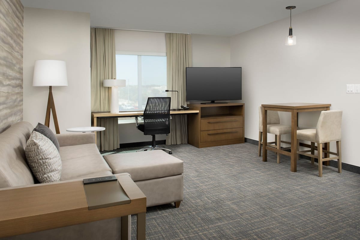 A hotel room with a sofa, a desk with a TV, a dining table with chairs, and a lamp.