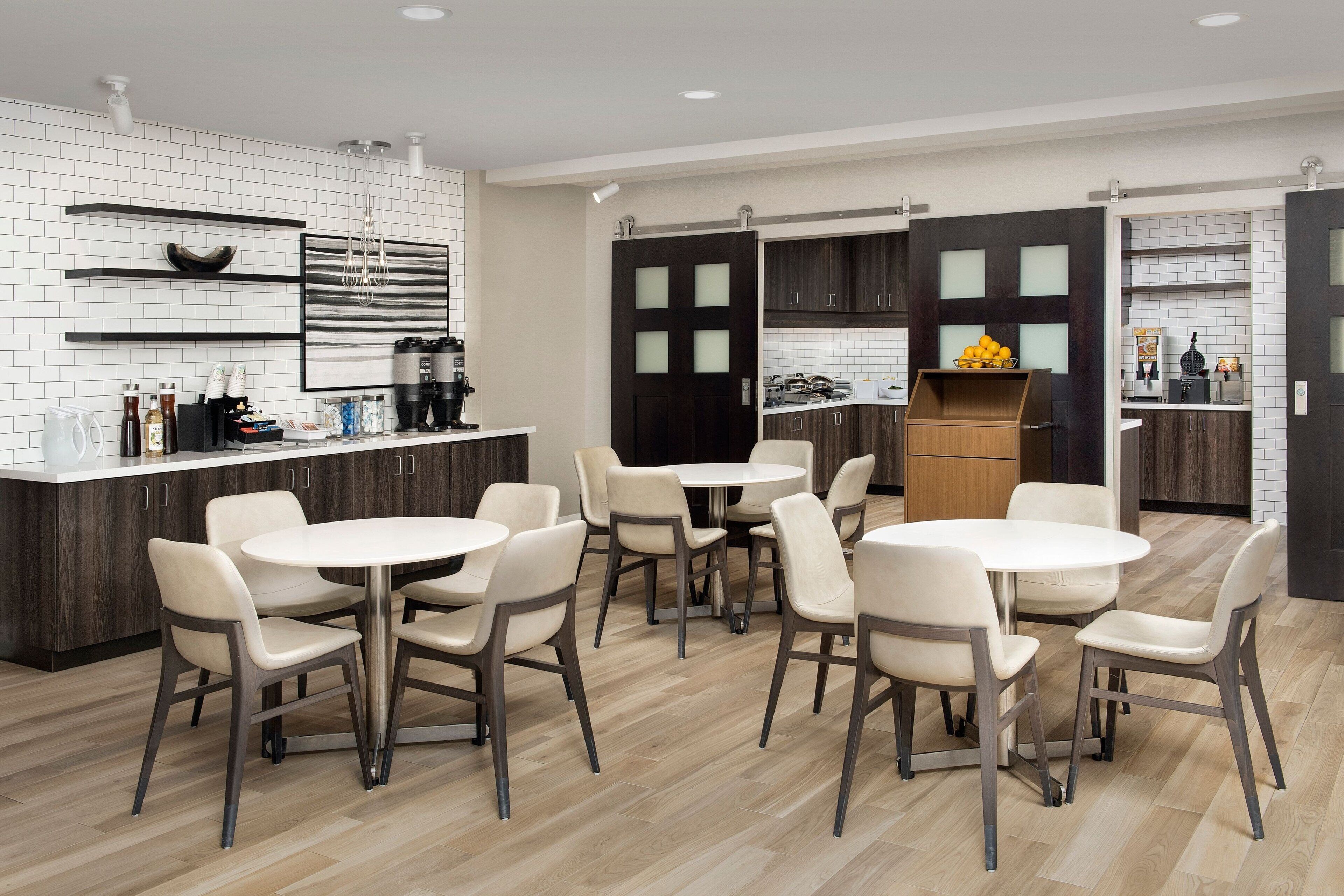 A modern kitchen with white and dark wood cabinetry, a central island with stools, and a dining area with round tables and chairs.