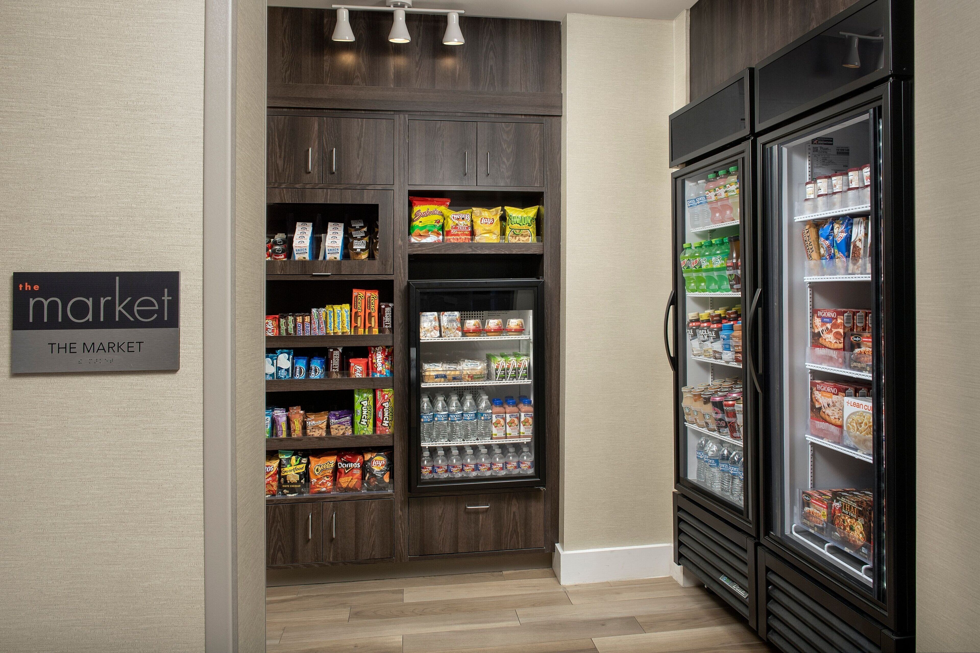Een kitchenette met een koelkast vol drankjes en snacks, een voorraadkast met diverse etenswaren, en een bordje met de tekst 'The Market'.