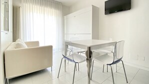 A modern, minimalist living room with a white sofa, a dining table with transparent chairs, and a flat-screen TV mounted on the wall.