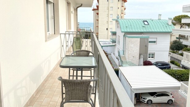 A balcony with a table and chairs, a car parked under a covered area, and a view of a building with a green roof.