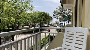 Un balcon avec une chaise blanche, un panneau de signalisation de type « stop », et une vue sur une rue bordée d’arbres et de bâtiments.