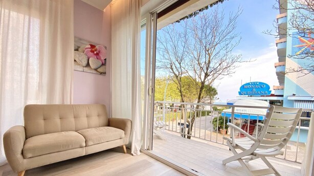 A balcony with a beige sofa and a white rocking chair, overlooking a building with a sign that reads "Monaco & Quisiana." - Appartamento Palladio 2 - Carraro Immobiliare Jesolo (Lido di Jesolo)