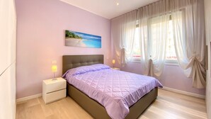 A bedroom with a bed, nightstands, a painting, and sheer curtains. - Appartamento Palladio 2 - Carraro Immobiliare Jesolo (Lido di Jesolo)