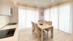 A modern kitchen with a built-in microwave, a sink, and a black dishwasher. There is a dining area with a wooden table and four chairs. The room has large windows with sheer curtains. - Appartamento Palladio 2 - Carraro Immobiliare Jesolo (Lido di Jesolo)