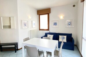 A dining area with a white table and chairs, a blue sofa, and framed pictures on the wall. - Appartamento Villa Santin 1 - Carraro Immobiliare Jesolo (Lido di Jesolo)