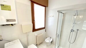A bathroom with a washing machine, toilet, bidet, and glass-enclosed shower.