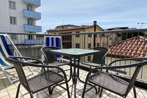 En balkon med et glasbord og fire fletstole, der ud til et boligområde med bygninger og en klar himmel.