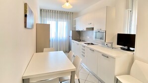 Une cuisine compacte avec des meubles blancs, une table à manger avec des chaises, un four encastré et un téléviseur posé sur le comptoir.