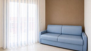 Une pièce avec un canapé bleu, une grande fenêtre ornée de rideaux légers et un mur sobre.