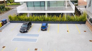 Un estacionamiento con dos autos, un edificio moderno con fachada de vidrio y una zona ajardinada con arbustos.