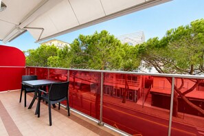 Un balcón con mesa y sillas, una pared roja y vistas a árboles y edificios.